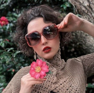 Woman wearing sunglasses and holding a pink flower, with a blurred natural background
