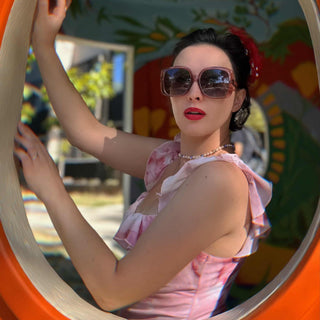 Woman wearing sunglasses and a pink dress standing inside a circular mirror with a colorful background.