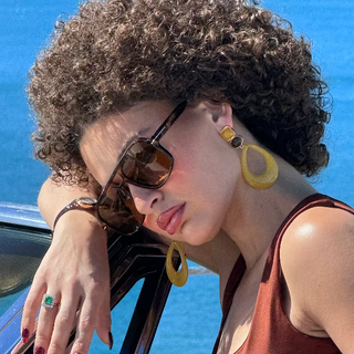 Woman with aviator sunglasses and earrings leaning on a car window against a blue ocean and sky.