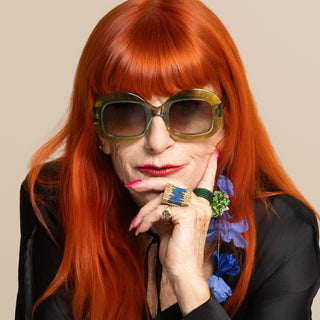 Person with red hair and green sunglasses posing with hand on chin against a beige background