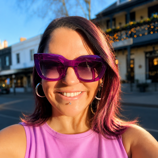 A smiling woman outdoors wearing oversized purple sunglasses and a bright pink sleeveless top, photographed in warm sunlight with a blurred street and storefront background.