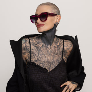 Person wearing a black outfit with intricate tattoo and sunglasses on a plain background