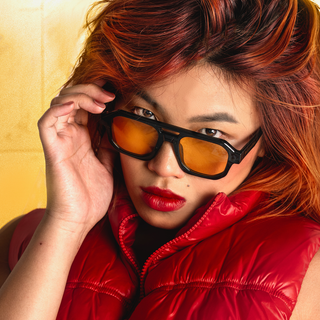 A close-up lifestyle shot of a model wearing thick black-framed sunglasses with yellow tinted lenses and a shiny red puffer vest, set against a textured gold background.