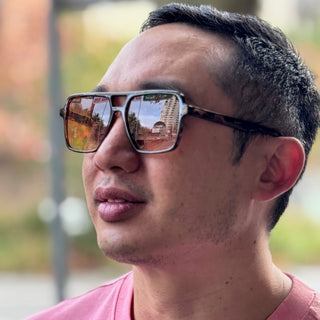 A close-up shot of a man wearing square aviator-style sunglasses with a tortoiseshell pattern on the temples and mirrored lenses reflecting an outdoor scene. 