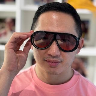 A man wearing oversized black aviator-style sunglasses with tinted lenses and a pink graphic t-shirt, holding the left side of the glasses with his hand.