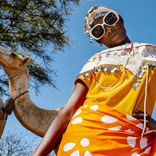 A woman wearing a bright yellow and orange patterned dress, a white beaded headwrap, and oversized white sunglasses, photographed outdoors against a blue sky with a camel visible behind her.