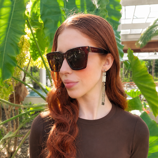 A woman with long wavy red hair wearing dark brown sunglasses and a brown long-sleeve top, standing outdoors in front of large green tropical leaves.