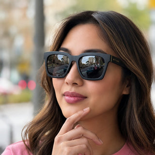 A woman with long brown hair wearing black square sunglasses and a pink t-shirt, posing outdoors with a blurred city street background.