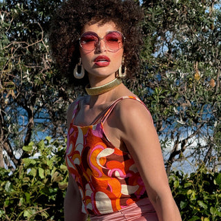 Woman in a colorful dress with pink round sunglasses and a gold necklace, standing outdoors with trees in the background.