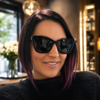 A woman with dark hair featuring purple highlights wears black cat-eye sunglasses and a black knit sweater, photographed in a warmly lit indoor setting.