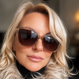 A close-up of a woman with blonde hair wearing oversized square sunglasses with a translucent brown frame and dark lenses, along with a fluffy beige faux-fur collar.