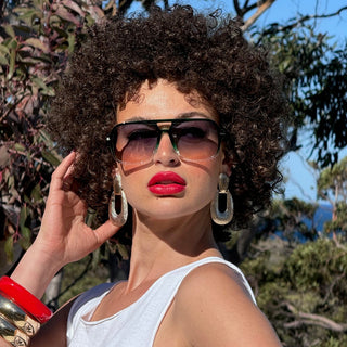 Woman with sunglasses and red lipstick outdoors, surrounded by greenery