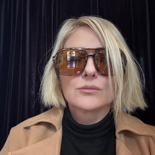 Person wearing sunglasses and a tan coat against a black background