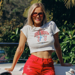 Woman wearing pink lens sunglasses and a white t-shirt with graphic design and red shorts, standing outdoors with greenery in the background.