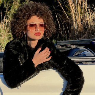 Person wearing a black fur coat and sunglasses, sitting in a car with a scenic background.