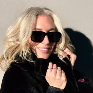 Person with blonde hair wearing sunglasses and a black coat against a neutral background