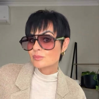 A person with short black hair wearing oversized aviator sunglasses with dark grey frames and pink-tinted lenses, a cream turtleneck, and a beige herringbone double-breasted blazer, standing indoors.