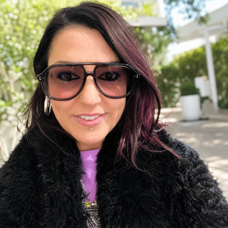 A close-up portrait of a woman wearing oversized grey aviator-style sunglasses, a black fuzzy jacket, and a purple top, photographed outdoors with blurred greenery and a patio in the background.