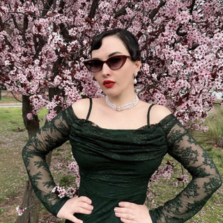 Woman in cat eye sunglasses and a green dress with sheer sleeves standing in front of cherry blossom trees.