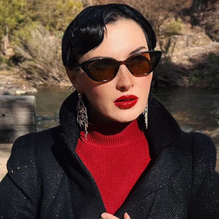 Woman wearing cat eye sunglasses, red lipstick, and a black coat with a red sweater against a natural background.