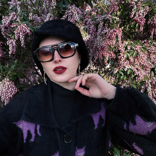 Person wearing sunglasses and a dark coat with a purple pattern, standing in front of pink flowering bushes.
