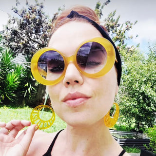 Person wearing large yellow sunglasses and matching earrings outdoors with greenery in the background