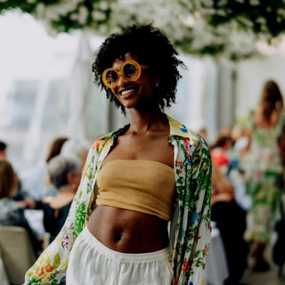 Woman wearing yellow sunglasses and a floral shirt in an outdoor setting with blurred people in the background