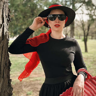 Woman in black outfit with red scarf and hat, standing outdoors near a tree.