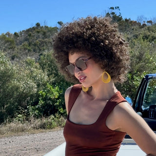 Woman with afro and brown sunglasses standing next to a car on a sunny day.