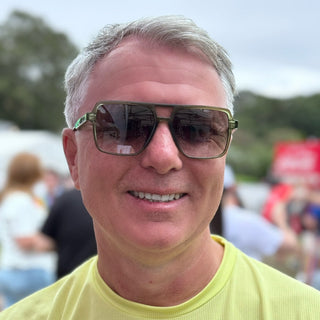 Man wearing sunglasses and a yellow shirt in an outdoor setting with blurred people and trees in the background
