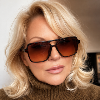 A close-up of a woman with blonde hair wearing oversized aviator sunglasses with dark brown lenses and tortoiseshell frames, paired with a brown turtleneck sweater.