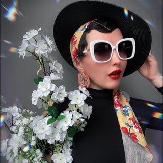 Person wearing a black hat and oversized white sunglasses, holding a vase of white flowers against a gray background.